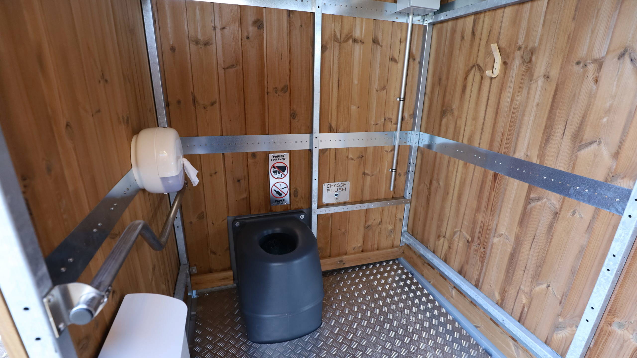 Intérieur de la cabine déplaçable bumper. La structure métallique est visible, constituée de tube en acier galvanisé. La cabine est habillée de bardage bois. Le sol est en tole alu gauffrée. La cuvette de couleur sombre est à gauche de l'image,. Sur la paroi gauche de la cabine, une barre pmr en forme de s allongé. Dessous une poubelle blanche. Au-dessus un distributeur de papier toilette de forme ronde,.